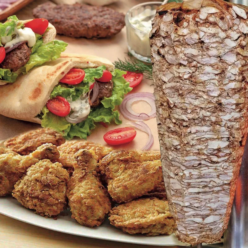 Gyros on pita with tomato and white sauce beside a platter of fried chicken and a gyro cone